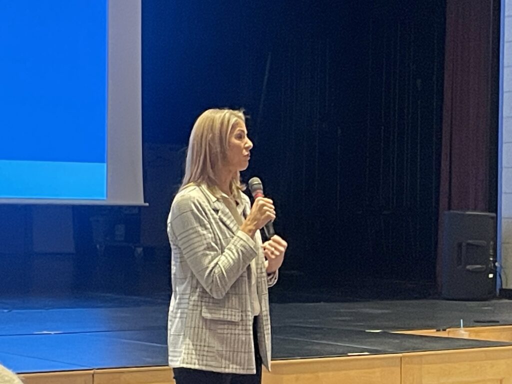 A tall blonde woman stands in front of a stage speaking into a microphone.