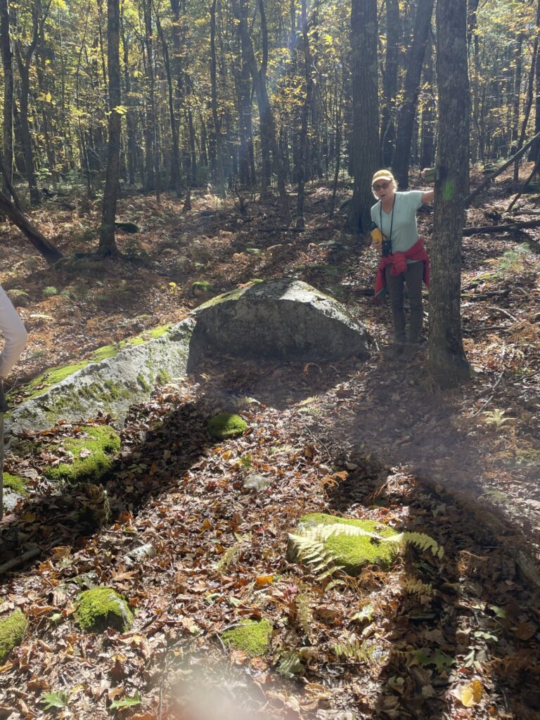 A woman (jacket now off on a warm day) stands b a tree next to a large boulder that seems to have been shaped by human hands.