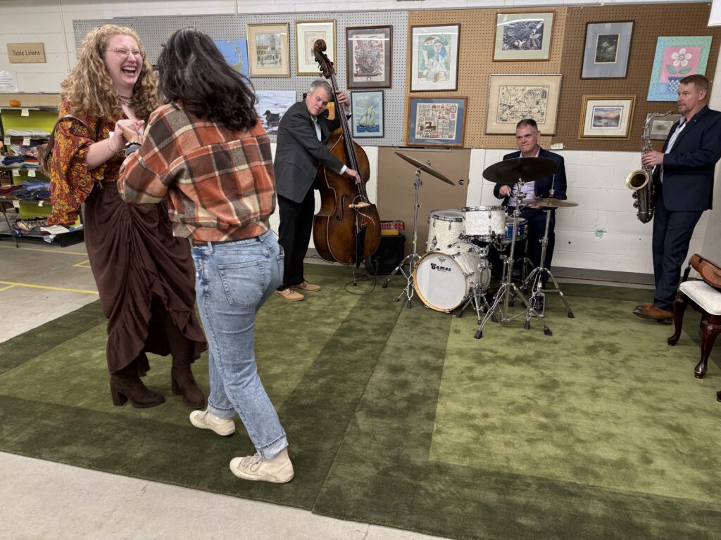 Two women, one wearing a skirt and the other wearing jeans, dance on a green carpet in front of a 3-piece band. The woman whose face is visible is laughing.