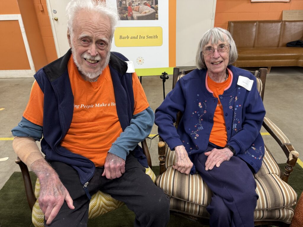 An older man and woman wearing orange Household Goods t-shirts sit on chairs in a warehouse.