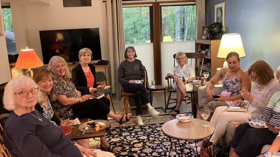 A group of women sit in a cozy looking living room. Some people have wine glasses and cheese plates.