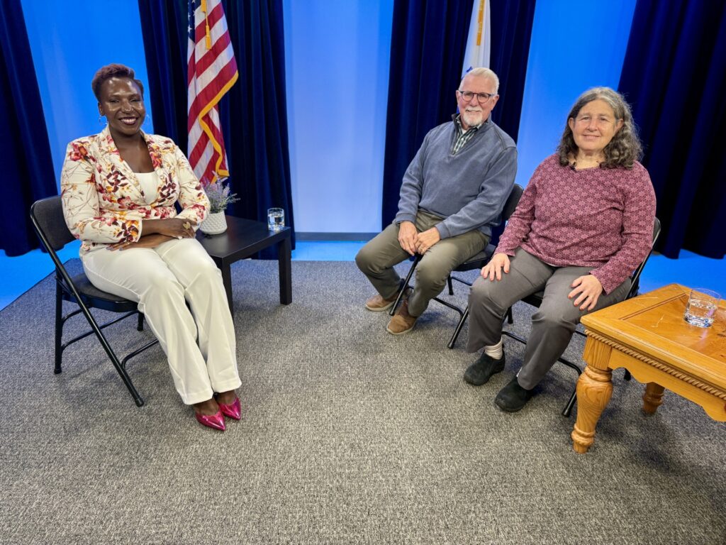Three people sit in a carpeted room. An American flag is in the background.
