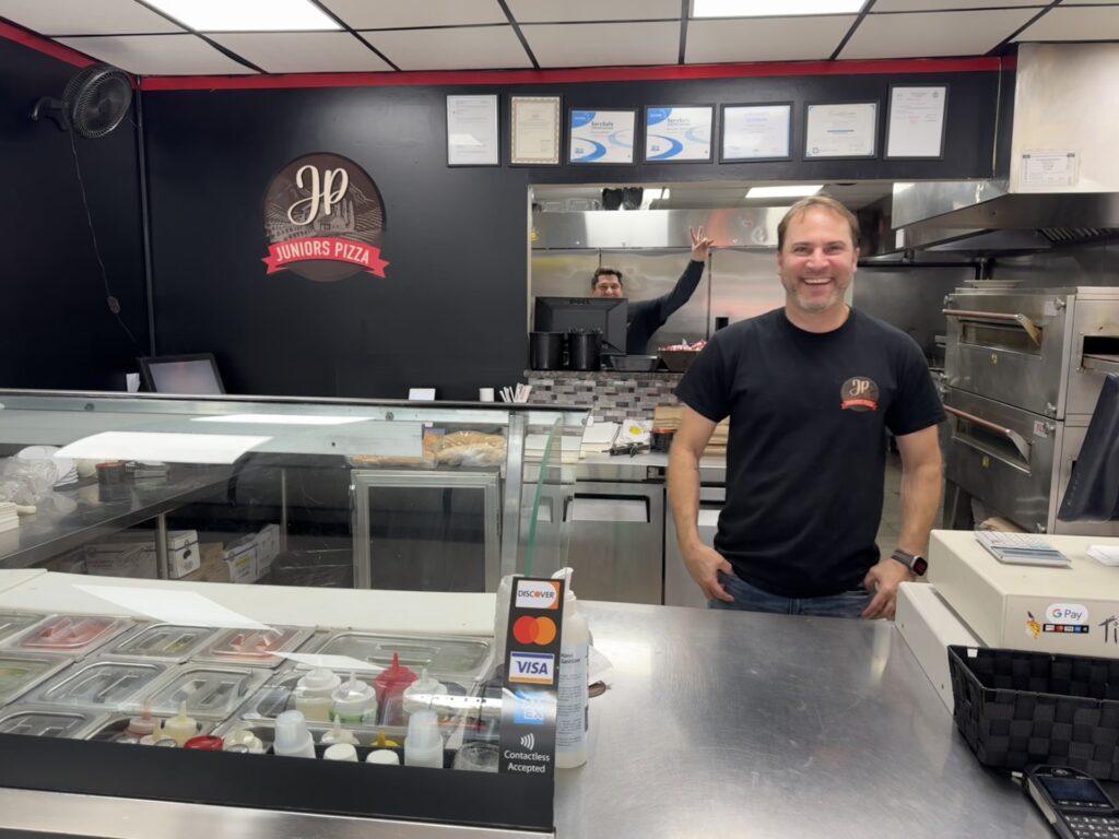A smiling man in a Junior's Pizza t-shirt stands behind a service counter. In the background, another man holds his hand up in a peace sign.