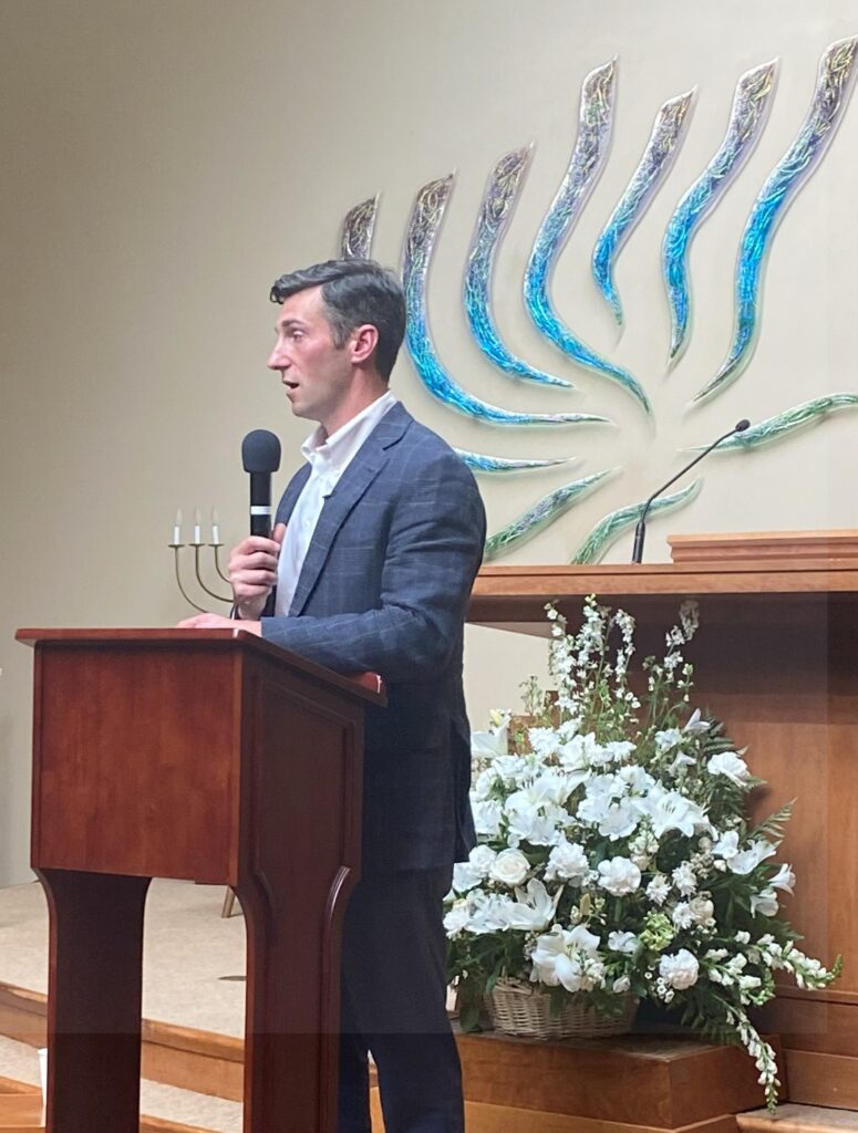 A man speaking from the bimah of a synagogue. In the background is an artistic representation of a menorah.