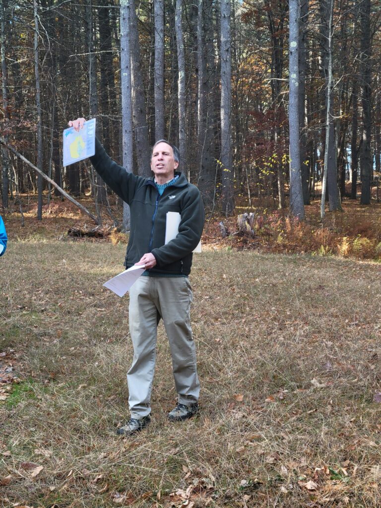 A man stands in a meadow in front of woods. He's holding up a map and speaking.