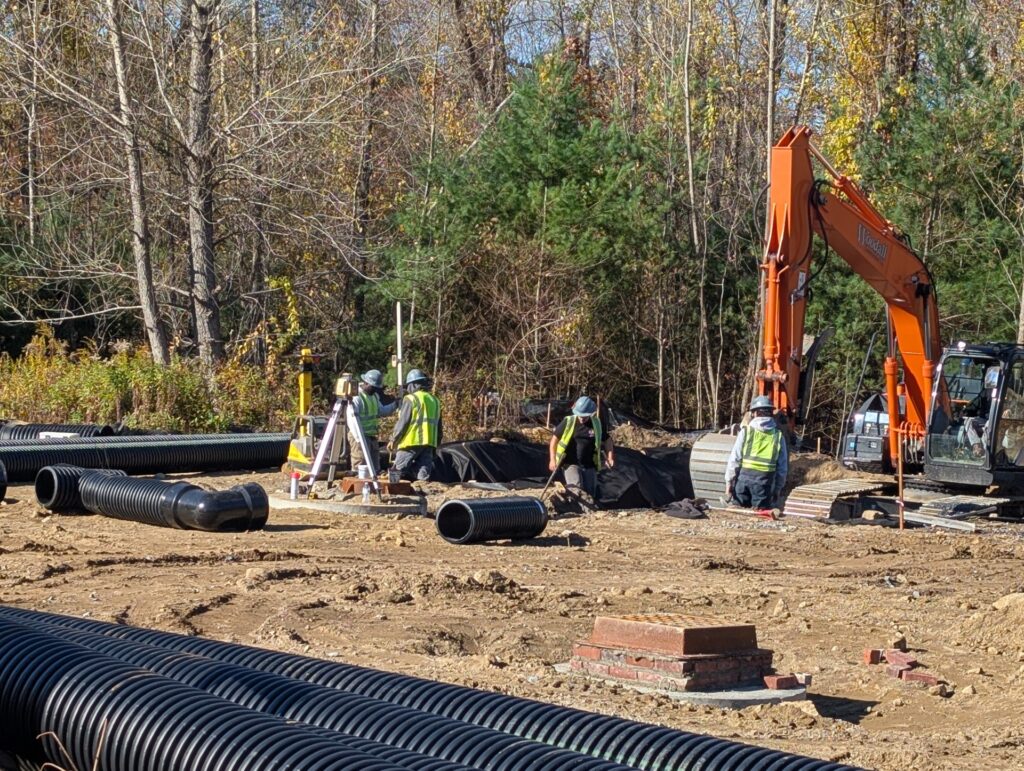 The foreground shows large black tubes. In the background is a large orange excavator and a bunch of people in hard hats and safety vests.