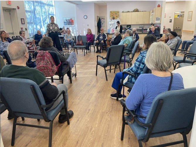 A group of people sit in chairs in a spacious room while a tall standing woman is speaking to them.