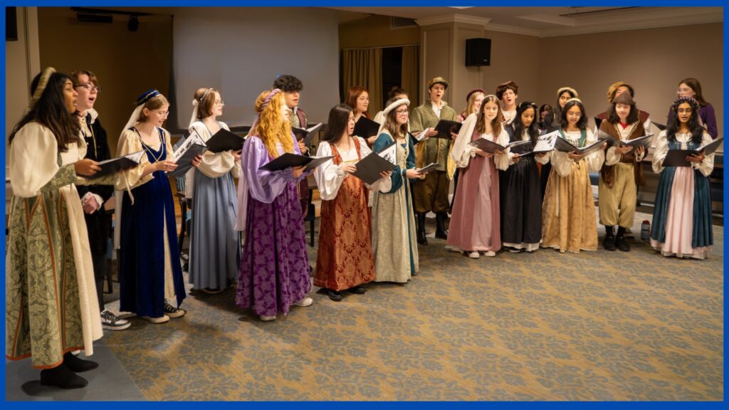 A group of singers wearing long dresses (for the women) and surcoats and puffy pants (for the men). Everyone wears great hats.
