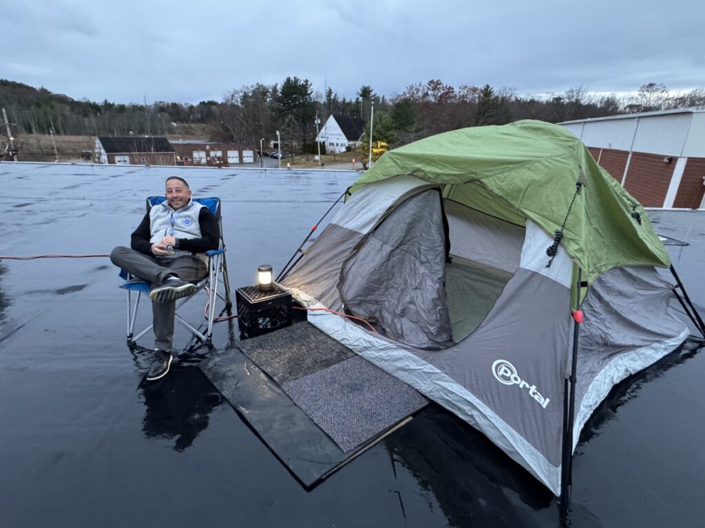 A man sits in a foldable chair on a rain-slicked roof. He has a small camping tent and a light ready to spend the evening.