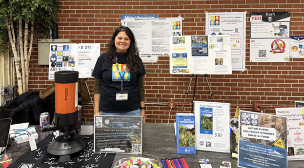 A woman wearing a STEM t-shirt stands behind a table full of information about various STEAM-related activities for youth.