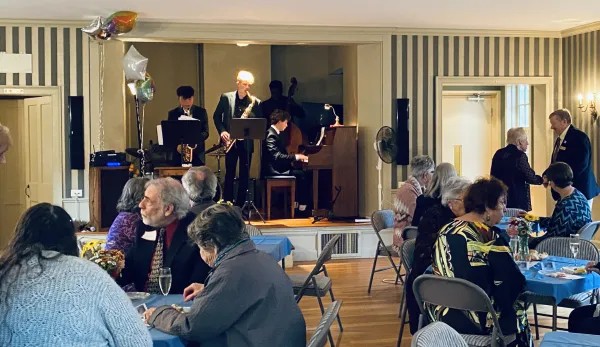 People seated at tables are chatting while in the background, four high-school musicians play music.