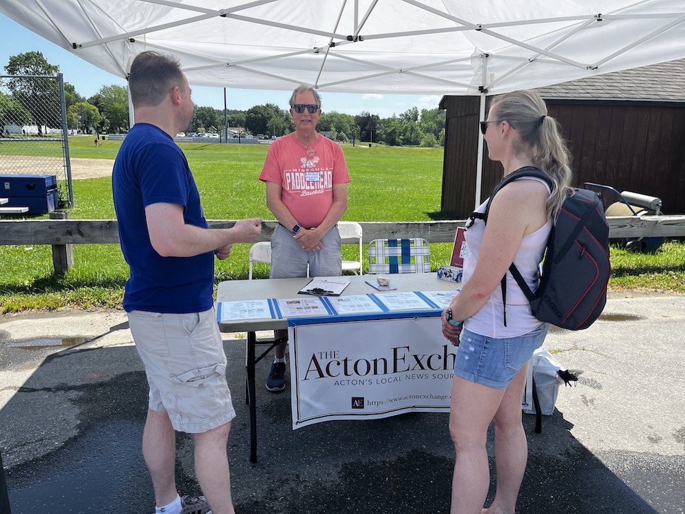A man and a woman in summer clothes chat with a man standing behind a table;  the table has a banner reading "Acton Exchange." 
