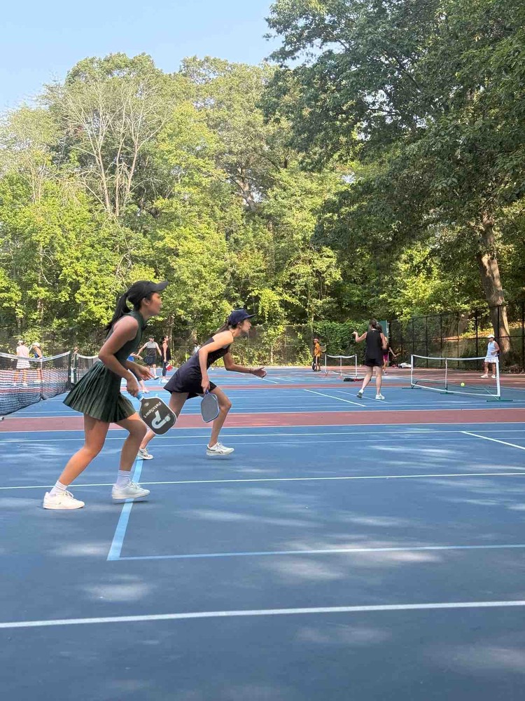 Two pony-tailed young women, dressed in black skirts, stand on a pickleball court wtih racquets ready to go. 