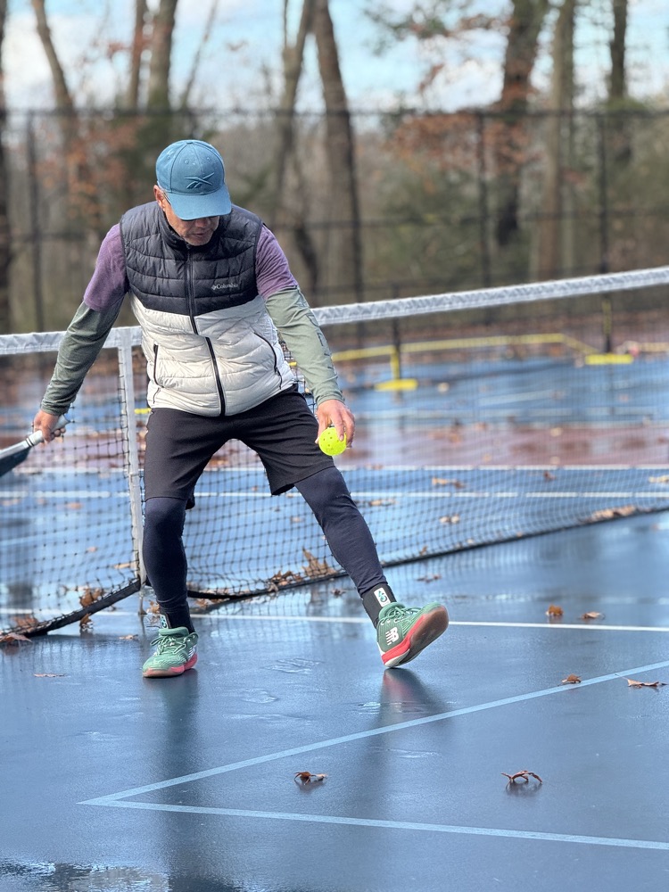 A man wearing long sleeves and a vest gets ready to hit a bright yellow ball from a rainy court.
