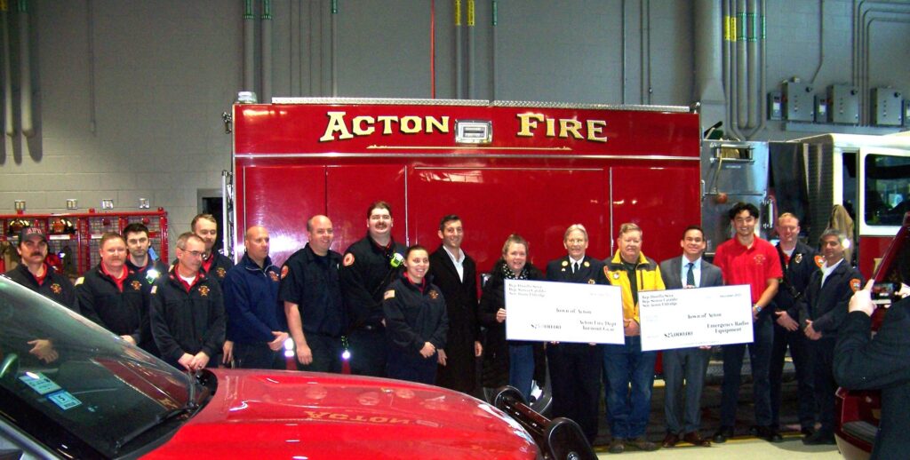 A group of people stand in front of a red Acton Fire truck. Several of the people are holding the two big checks.
