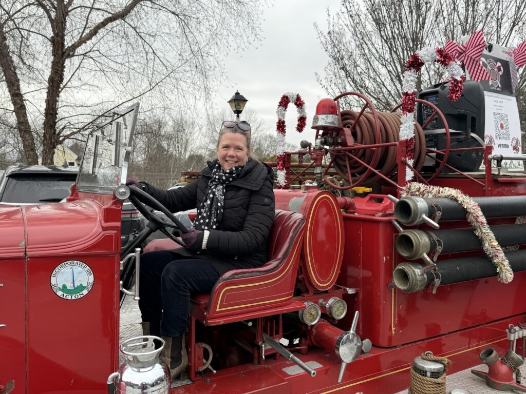 A grinning woman sits atop an antique fire truck with her hands on the steering wheel.