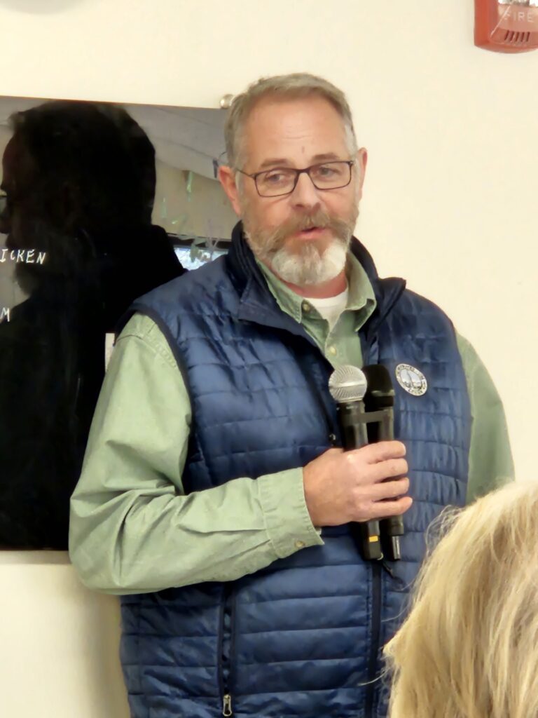 A bearded man with glasses, wearing an Acton vest, speaks into a double mic.