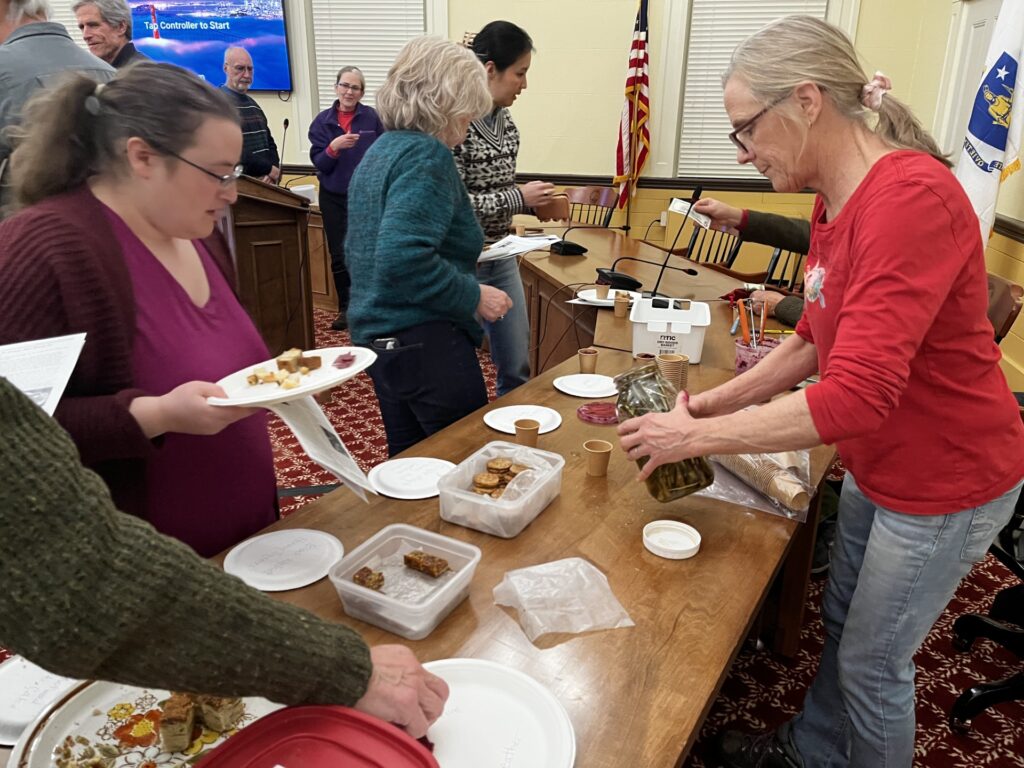 People line up at a table with snacks. A woman in a red shirt pours something out of a jug.