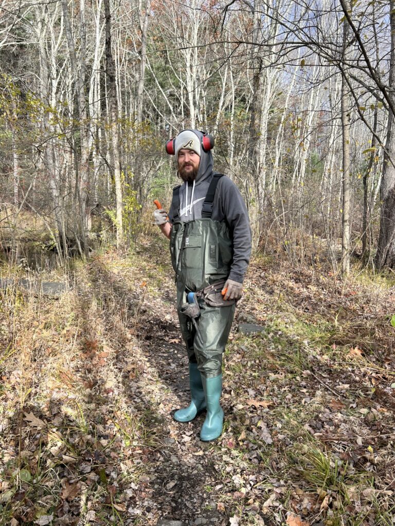 A bearded man wearing full chest waders, rubber gloves, a wool hat, and ear protectors.