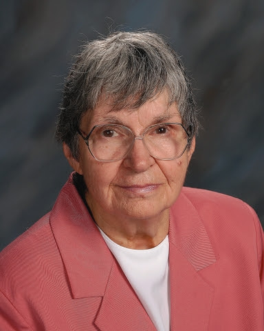 A woman with short grey hair and glasses wearing a pinkish jacket.