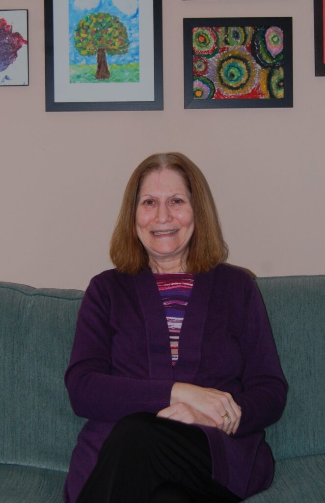 A woman wearing purple sits on an aqua couch with several works of art in the background.