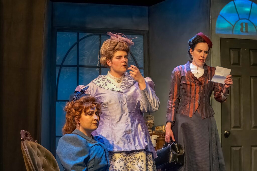Three women in Victorian dress are on a stage. The woman in the middle, wearing a lilac jacket, is smoking a pipe. The woman on the right (Dr. Watson), carries a leather medical bag.