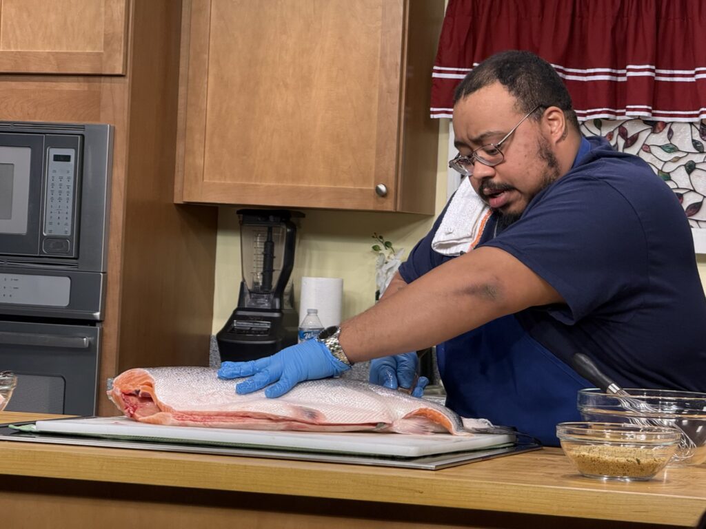 A man with glasses, gloves, and Twin Seafood T-shirt tackles a large chunk of fresh looking salmon.