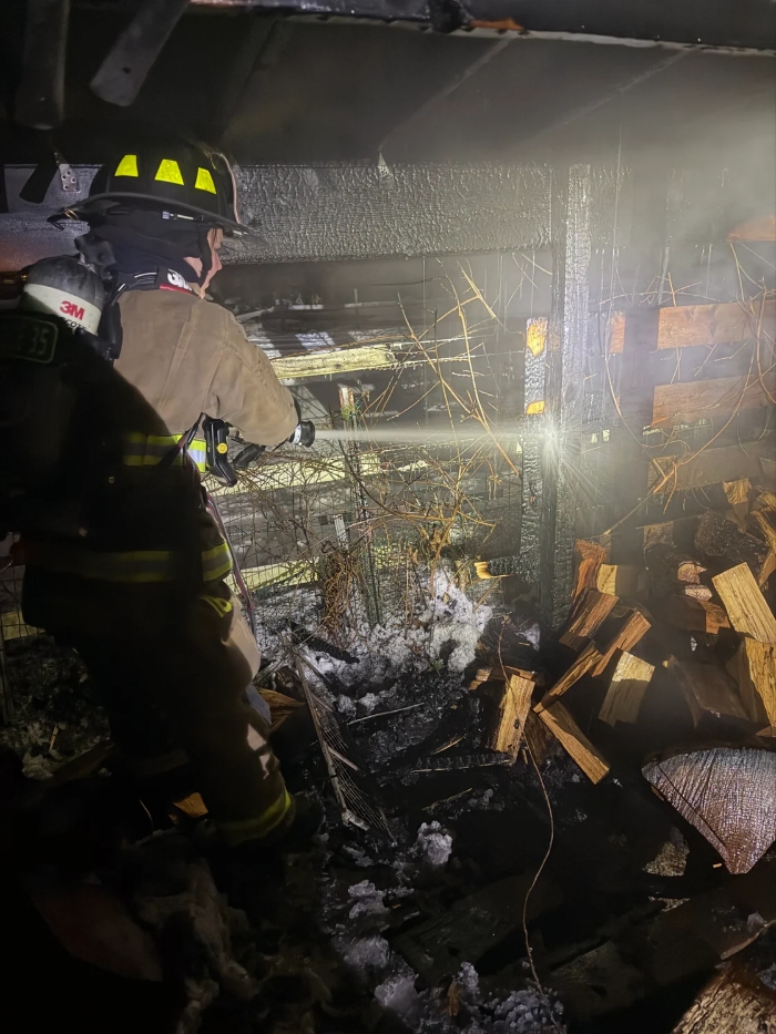 A firefighter aims a hose at charred sections of wood. There are stacks of firewood and other unidentifiable objects in the picture as well.