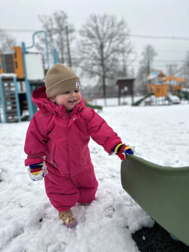 A very small human in a very pink snowsuit holds on the the bottom of the slide at a playground.