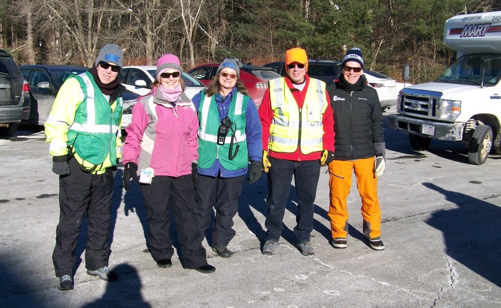 A group of warmly dressed people, some wearing vests, stands in the sun.