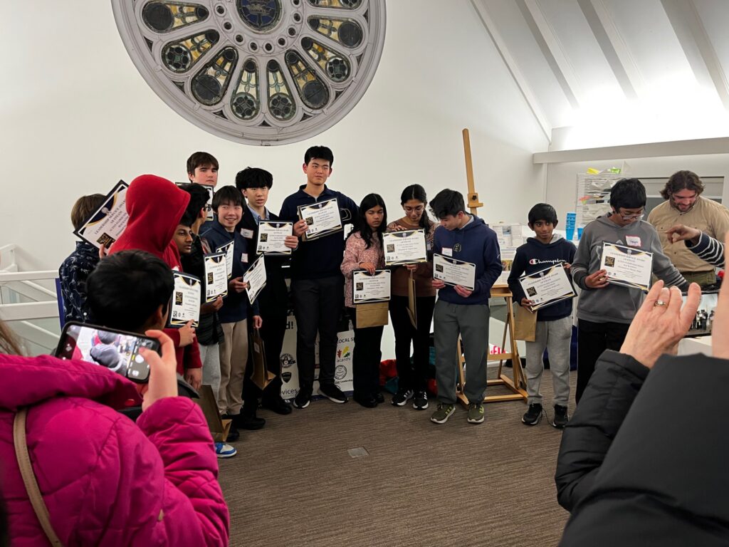 Fourteen youth display certificates. Onlooking adults take photographs with cell phones.