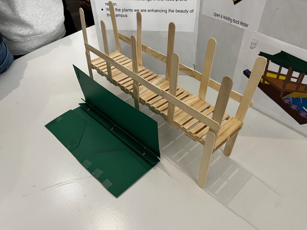 Student-made model of a bridge with a dam protection device in front of it. The model bridge is made of popsicle sticks and the model dam protection device is made from a folder, open to show how one limb of the device stays on the streambed while the other limb rises up to protect the bridge.