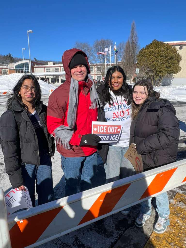 Three high school aged women stand posing for the photo with a taller man. All are bundled up against the cold, but smiling. The man holds a sign that says "ICE OUT" in English and Spanish.