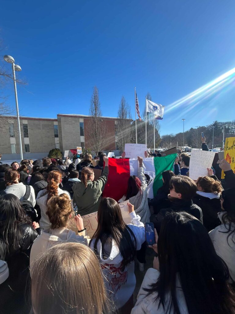 A crowd of high school aged youth are seen from behind, apparently intent on a speaker in the far distance. Several carry flags or signs.