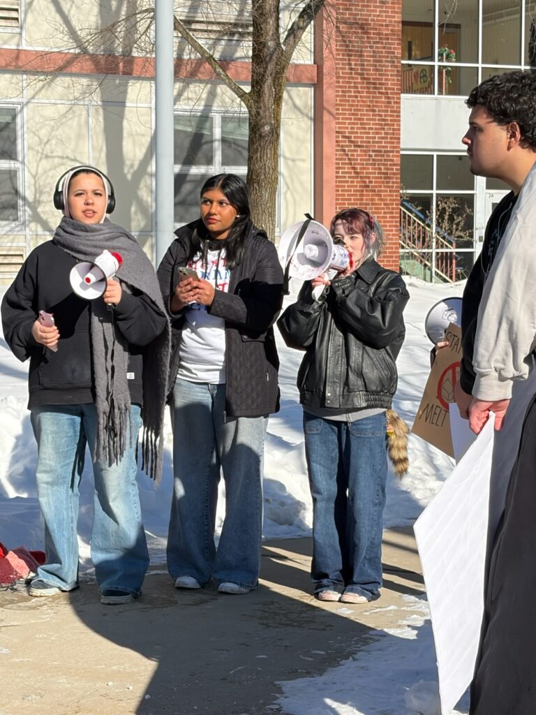 With piles of snow and a school building visible in the background, three high school aged youth address a gathering. Two of the speakers hold hand-held, battery-powered megaphones.