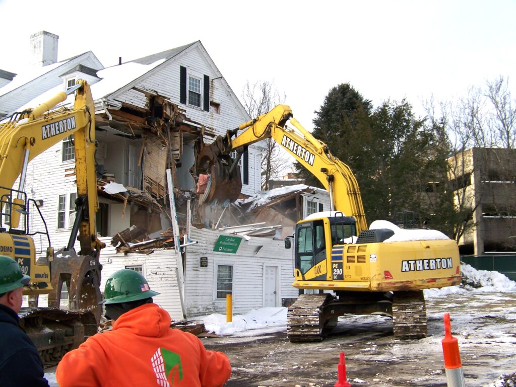 Two large yellow excavators start demolishing a white, wood-frame house.