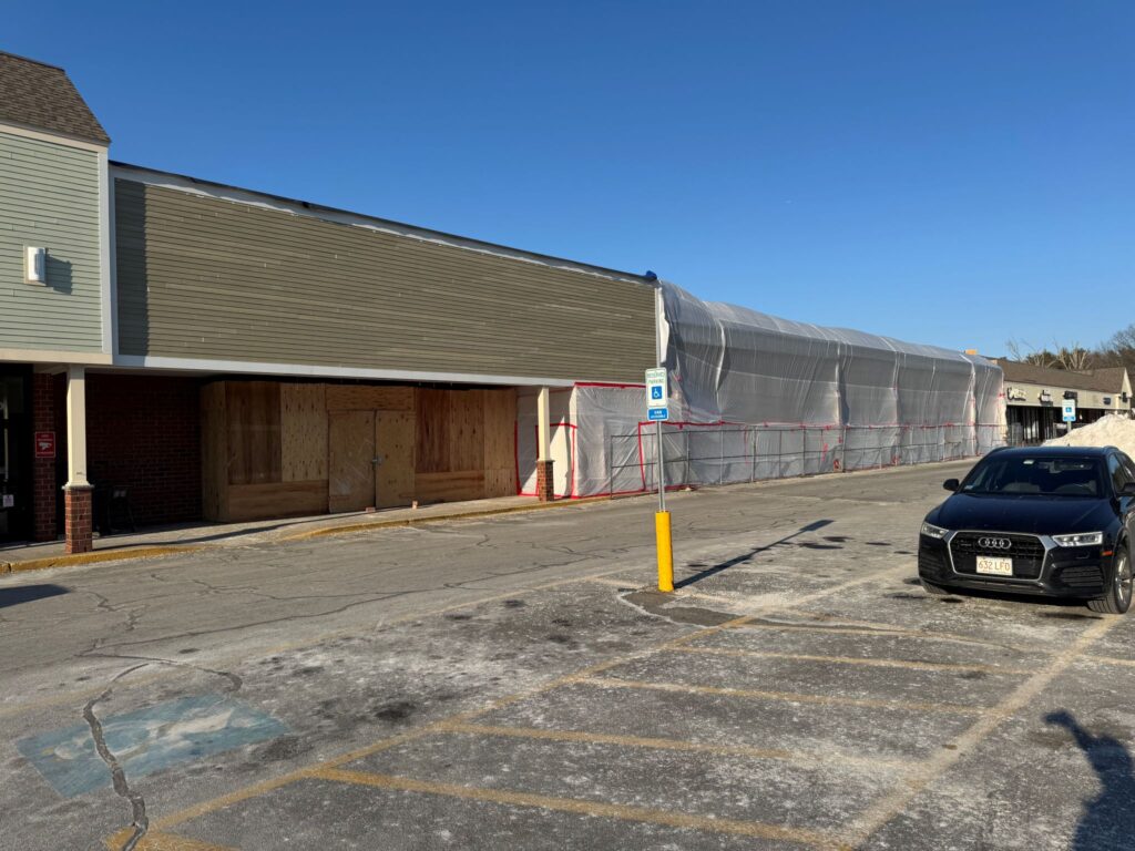 A mostly empty parking lot and a storefront divided in two. The left side is open and has a lot of plywood at the entrance. The right sighed is completely covered with large sheets of plastic.
