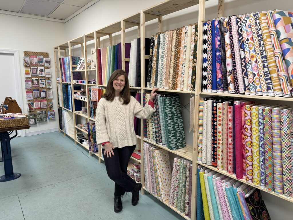 A smiling woman stands in front of shelves full of multicolored bolts of fabric. On the back wall are quilt patterns and interesting sewing notions.