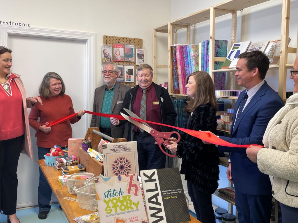 A line of people holding a red ribbon in a store. A woman is cutting the ribbon with oversized scissors.