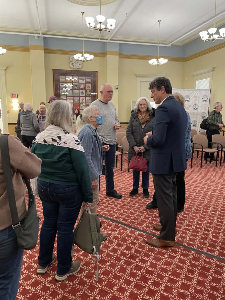 A number of people standing around Massa Viana. On the left, more people are standing in line to talk to him, and in the background other gorups of people are chatting.