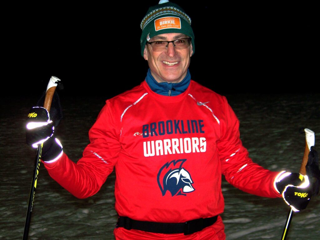A smiling man wearing a Brookline Warriors jersey, ski gloves, and a hat with a bobble stands with his ski poles.