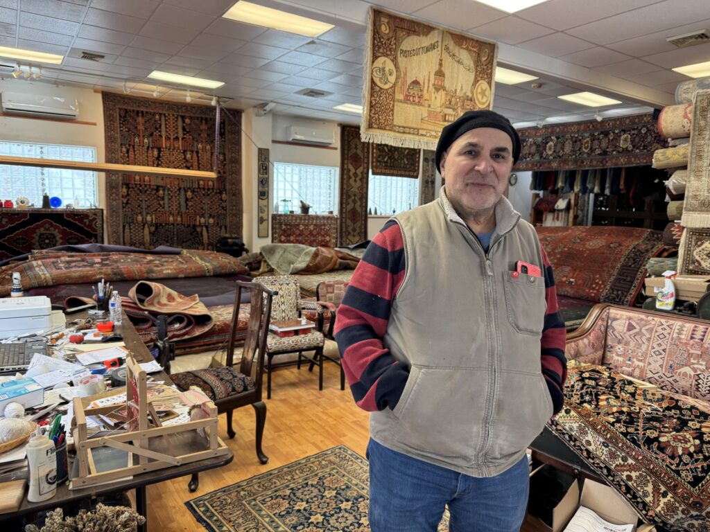A man wearing a beret and a vest stands in a store filled with carpets.