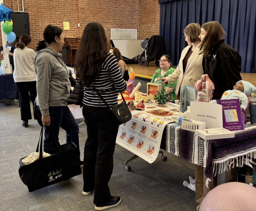 Two people are standing at a table full of information and books. Several people behind the table are chatting with the guests. One person carries a large bag that says "oh baby!" on the side.
