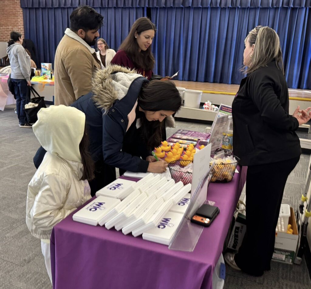 Four people, including a child, stand in front of a table that is covered with interesting items, including white plastic boxes that say WIC and a fleet of yellow rubber ducks. A woman behind the table is chatting with one of the people.