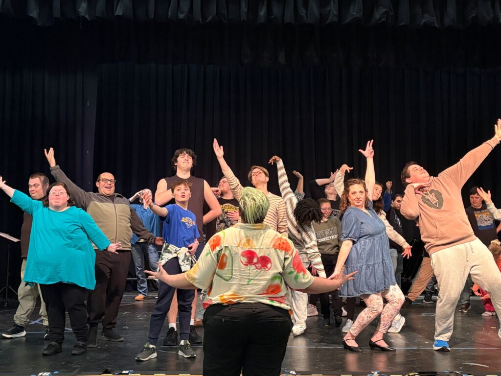 Disabled and nondisabled actors sing while striking poses while the choregrapher with green hair stands at the edge of the stage wearing a shirt with colorful fruit.