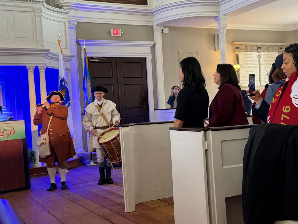 Two people wearing colonial dress and tri-corner hats stand at the front of a building. The woman on the left is playing a fife, the man on right is beating a drum.