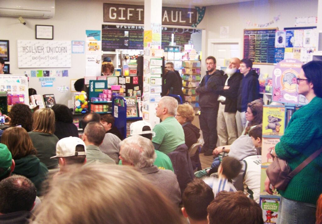 A very croweded small space with every seat filled and people lined up against the bookcases in the back.