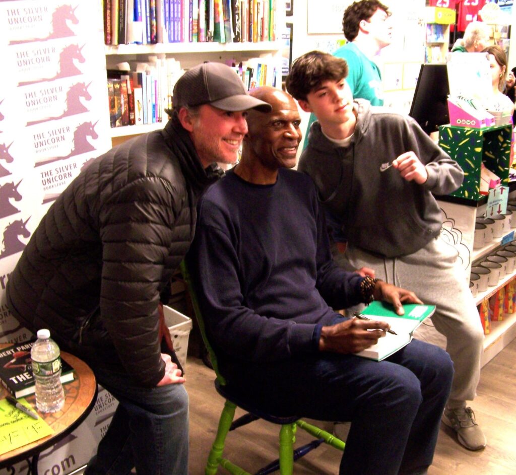 A very tall seated Black man in blue sweats pauses signing a book while two people (probably a dad and preteen son) pose with him.