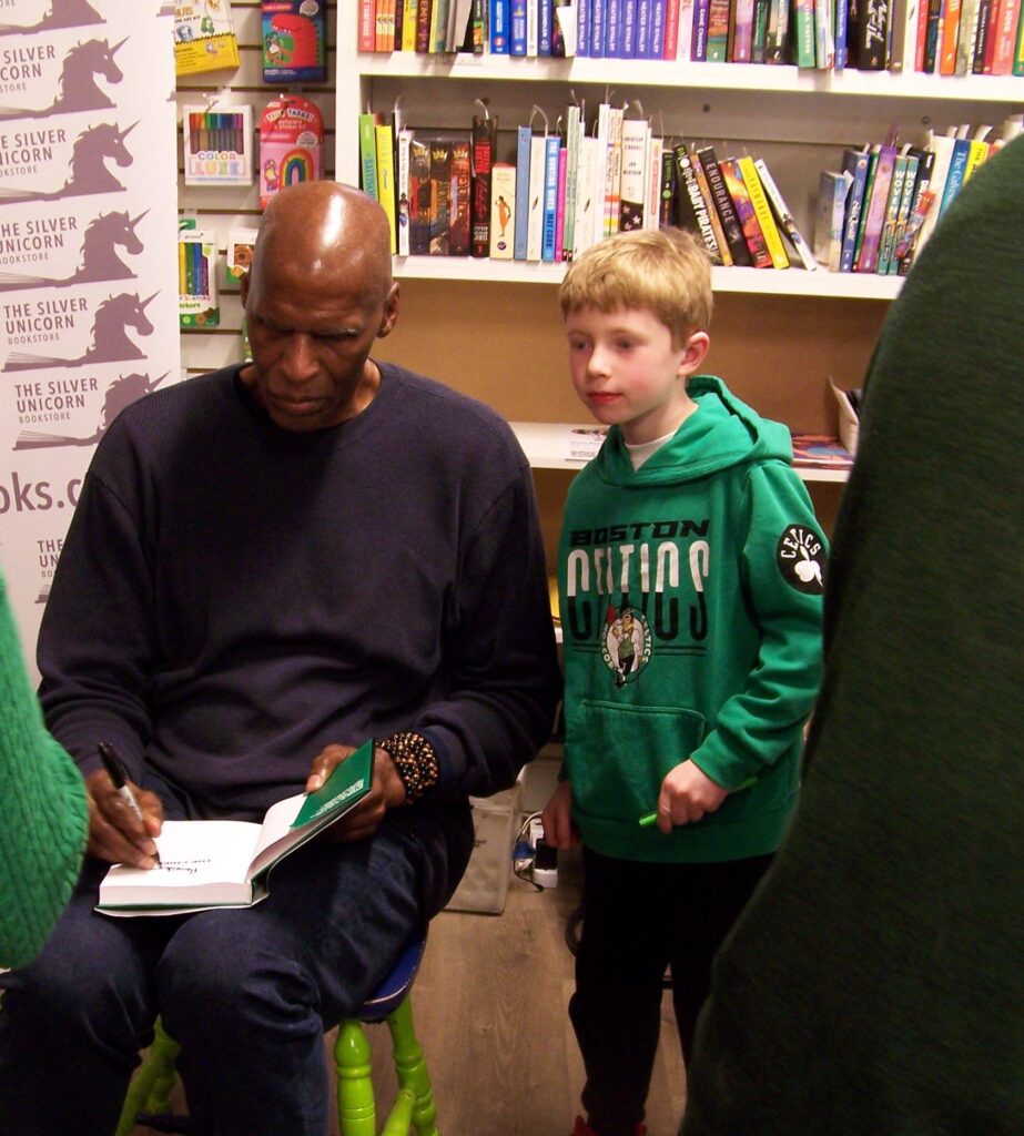 A tall, bald, Black man signs a book in his lap while a young boy looks over his sholder.