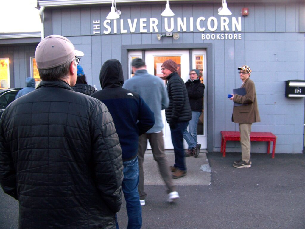 A gray building with a sign that says Thye Silver Unicorn Bookstore. Heading into the building is a long line of people, mostly men.